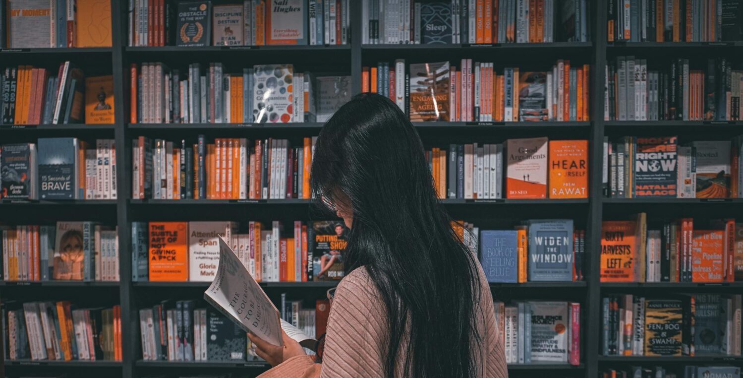 Young woman engrossed in a book amidst colorful bookstore shelves.