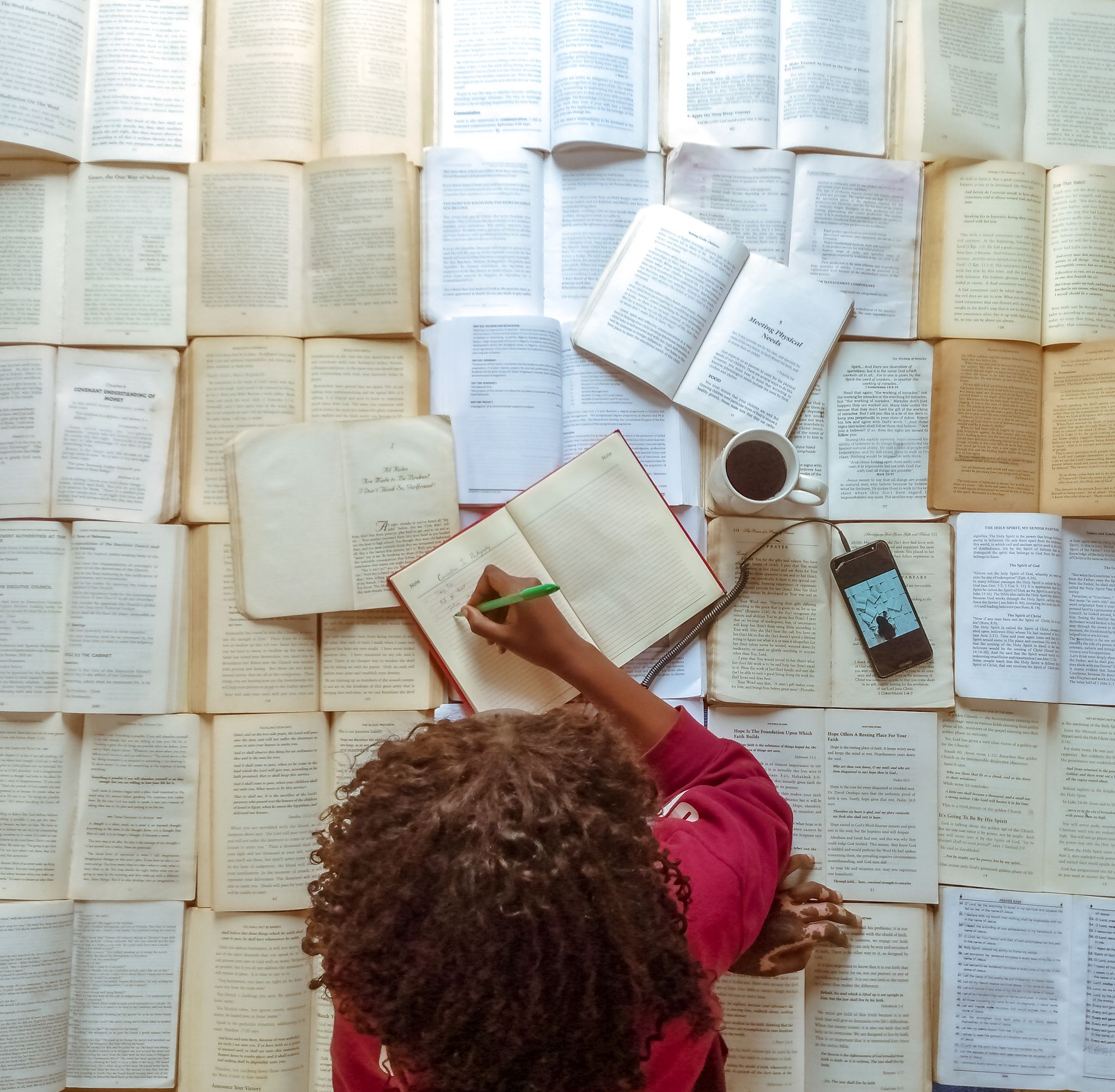 A woman writes in a notebook surrounded by open books with a cup of coffee nearby.